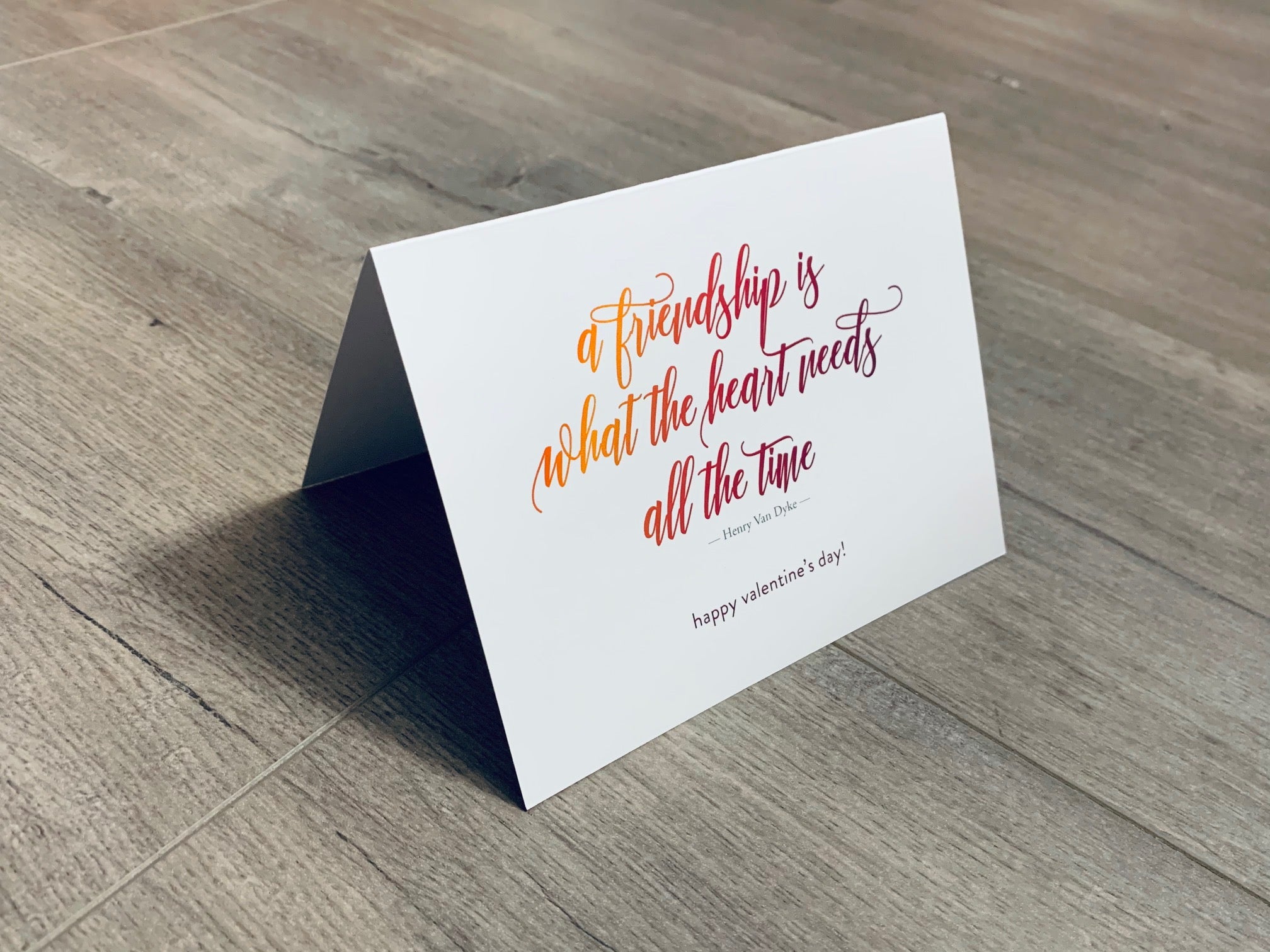 A white, folded notecard sits on a wooden floor. The card has a quote by Henry Van Dyke and says, "a friendship is what the heart needs all the time." The Friendship Valentines collection by Stationare.