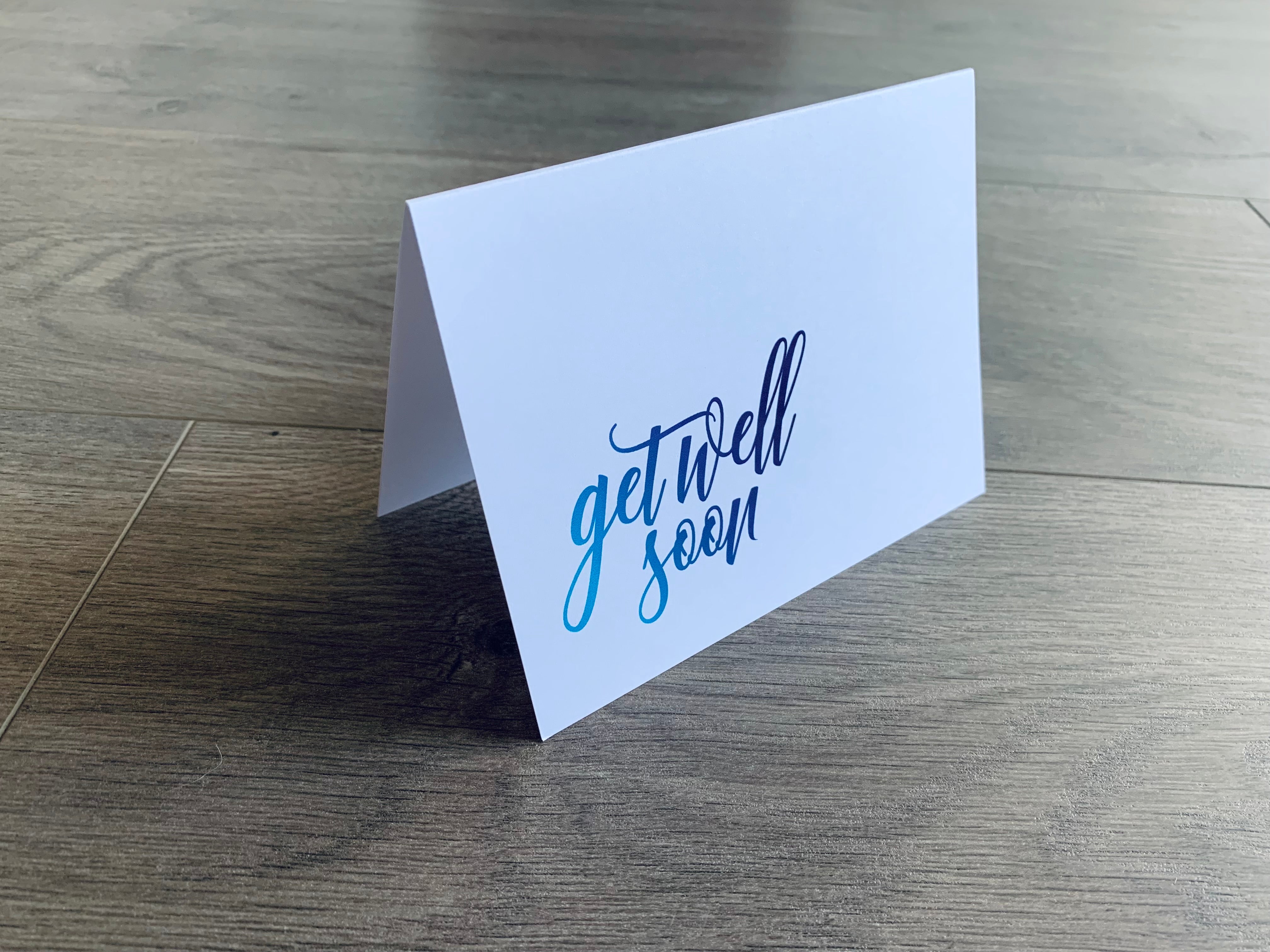 A white folded card is propped up on a gray wooden background. The card reads, "Get Well Soon" in the bottom corner in a script font that fades from light blue to navy. From Stationare's Get Well Soon Collection.
