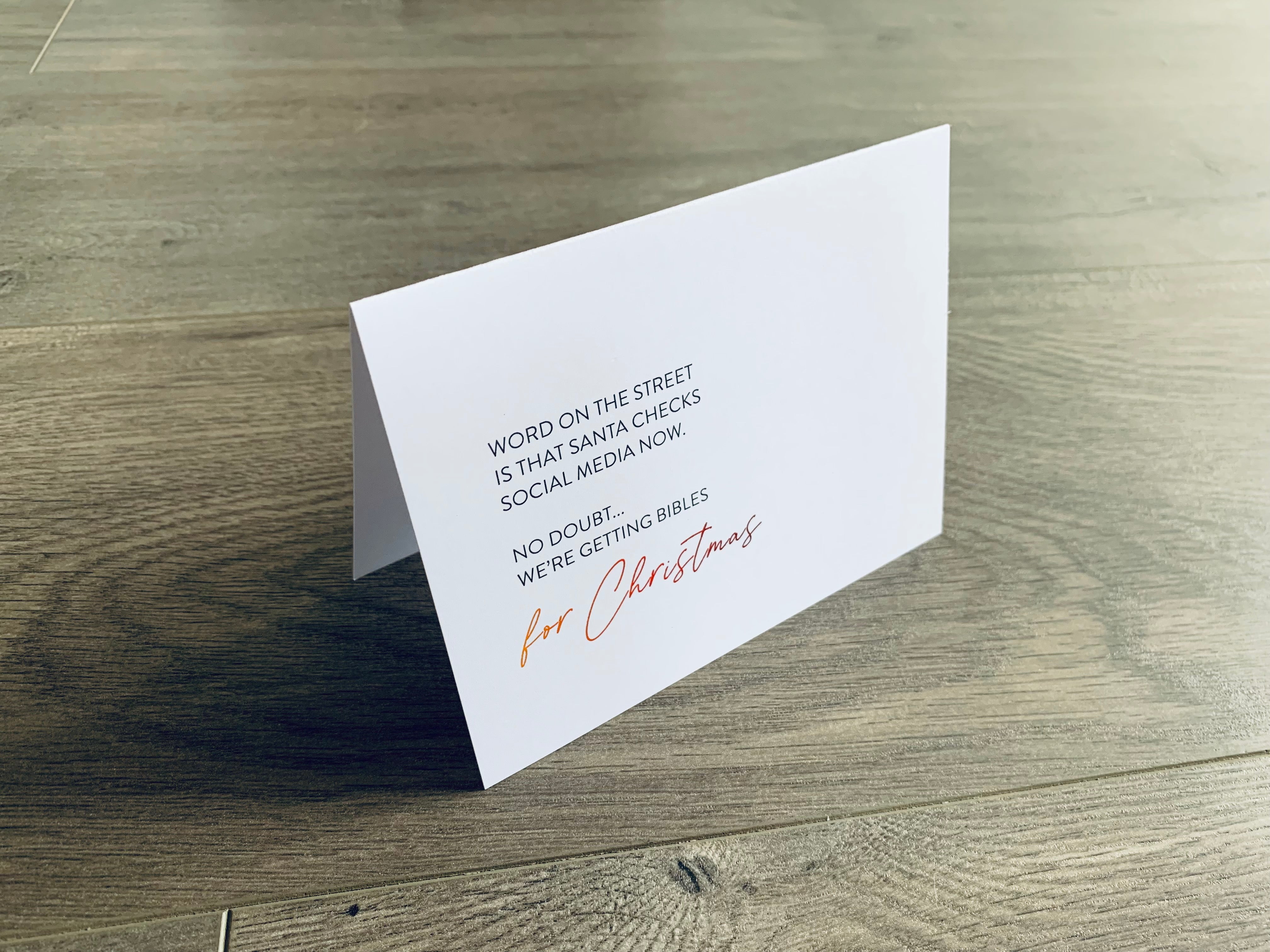 A white folded notecard is propped up on a wooden floor. On the front of the card, it says "Word on the street is that Santa checks social media now. No doubt... we're getting bibles for Christmas." Christmas Chuckles collection by Stationare.