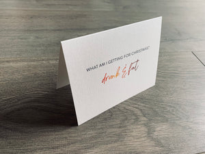A folded notecard is propped up on a gray wood floor. The notecard is of a cream-colored pearlized paper. The card says, "What am I getting for Christmas? drunk and fat" Christmas Chuckles collection by Stationare.