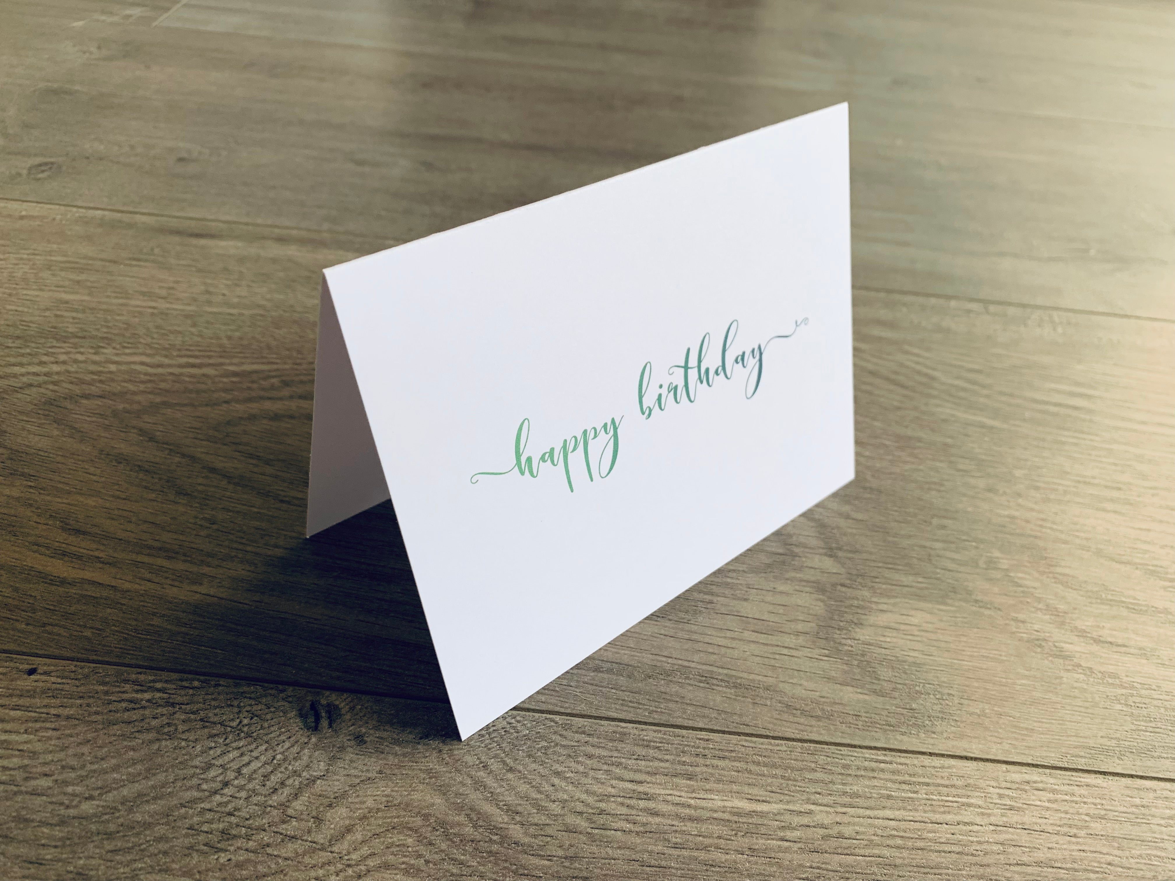 A single white notecard is propped up on a wooden floor. The card reads, "happy birthday" in a fancy script font in green ombre. Happy Birthday Collection by Stationare.