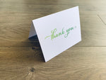 A white notecard with a script font that reads "thank you" sits on a wooden floor. The font fades from light green to dark green.