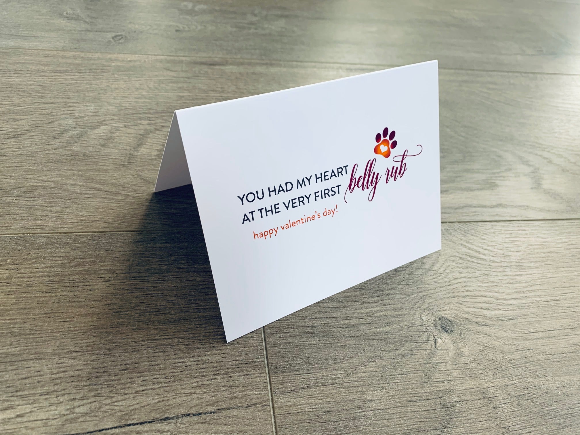 A folded white notecard is folded and propped up on a gray wooden background. The card says, "You had my heart at the very first belly rub. Happy Valentine's Day!" Valentines from the Pup collection by Stationare.