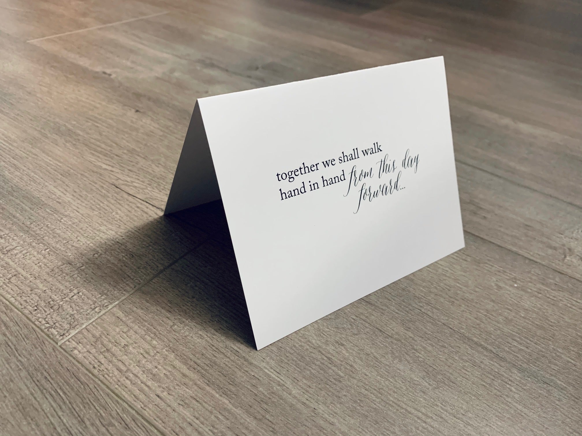 A white, folded notecard is propped up on a gray wooden floor. The card says, "together we shall walk hand in hand from this day forward..." in black ink. Stationare's Bridal and Wedding collection.