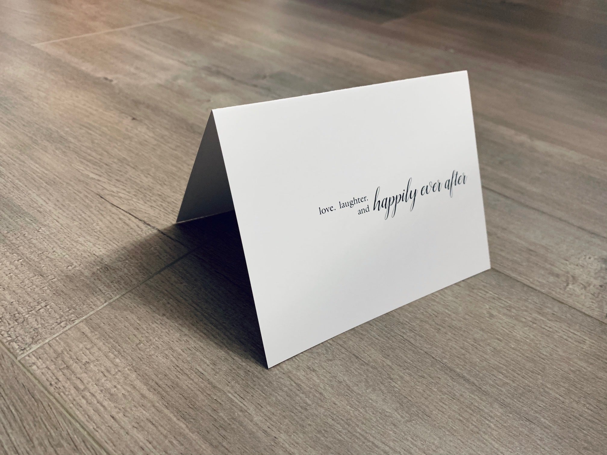 A white card that reads, "love, laughter and happily ever after" in black ink is folded and propped up on a wooden floor. Bridal and wedding card from Stationare.