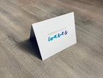 A white card is propped on a gray wood floor. The card reads, "Happiness comes in waves." Beach Life Collection by Stationare.