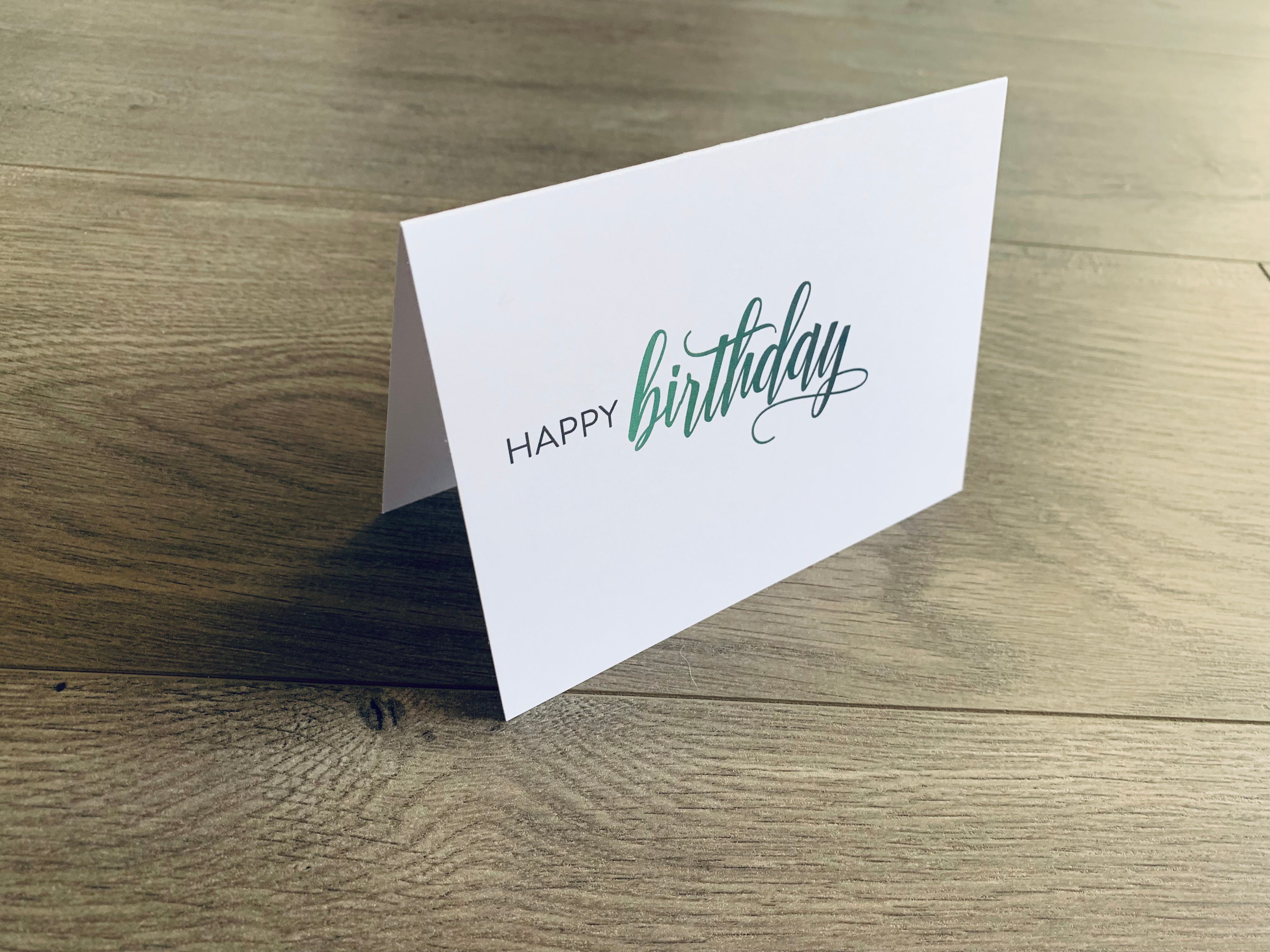 A single white notecard is propped up on a wooden floor. The card reads, "happy birthday" in a block and script font. Happy Birthday Collection by Stationare.