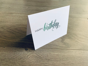 A single white notecard is propped up on a wooden floor. The card reads, "happy birthday" in a block and script font. Happy Birthday Collection by Stationare.
