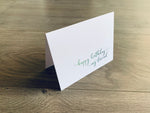 White folded notecard is propped up on a wooden floor. The card reads, "happy birthday, my friend" in a script font. From Stationare's Friendship Birthday collection.
