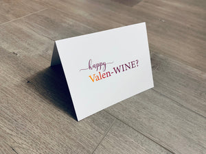 A folded white notecard is folded and propped up on a gray wooden background. The card says, "happy valen-wine?" Valentine Smiles collection by Stationare.