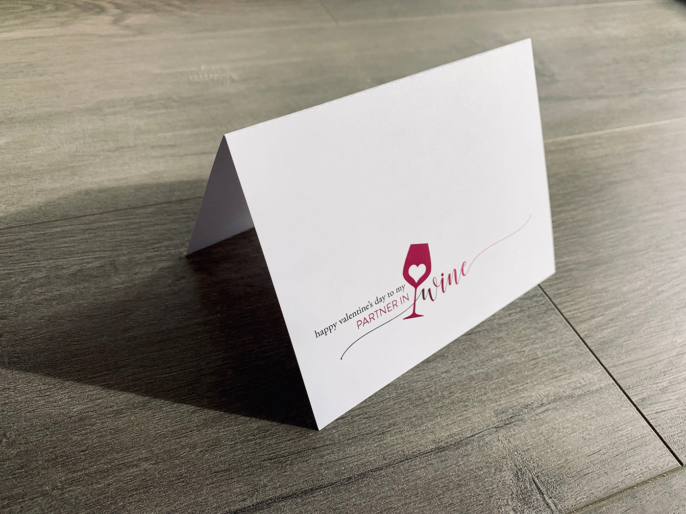 A folded white notecard is propped up on a wooden floor. The card says, "Happy Valentine's Day to my partner in wine" with a heart inside a wine glass. Stationare's Valen-wine collection.