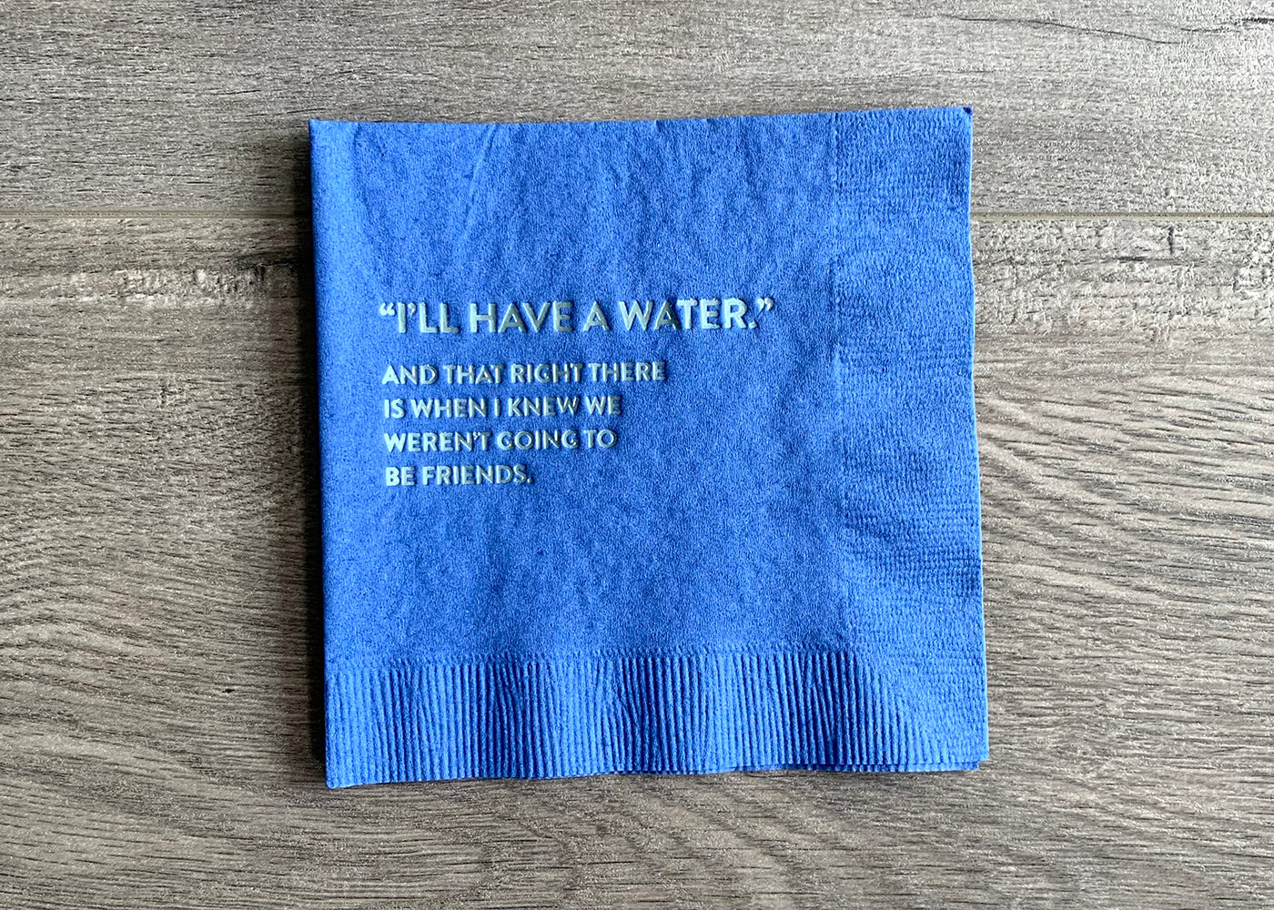 A royal blue cocktail napkin lies on a wooden background. In metallic light blue foil, it reads a quote "I'll have a water." with the wording "and that right there is when I knew we weren't going to be friends."