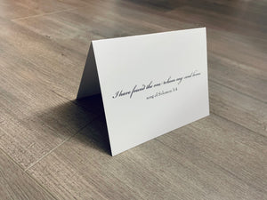 A folded white card is propped up on a wooden floor. In black ink it reads, "I have found the one whom my soul loves. Song of Solomon 3:4" Bridal and Wedding collection by Stationare.