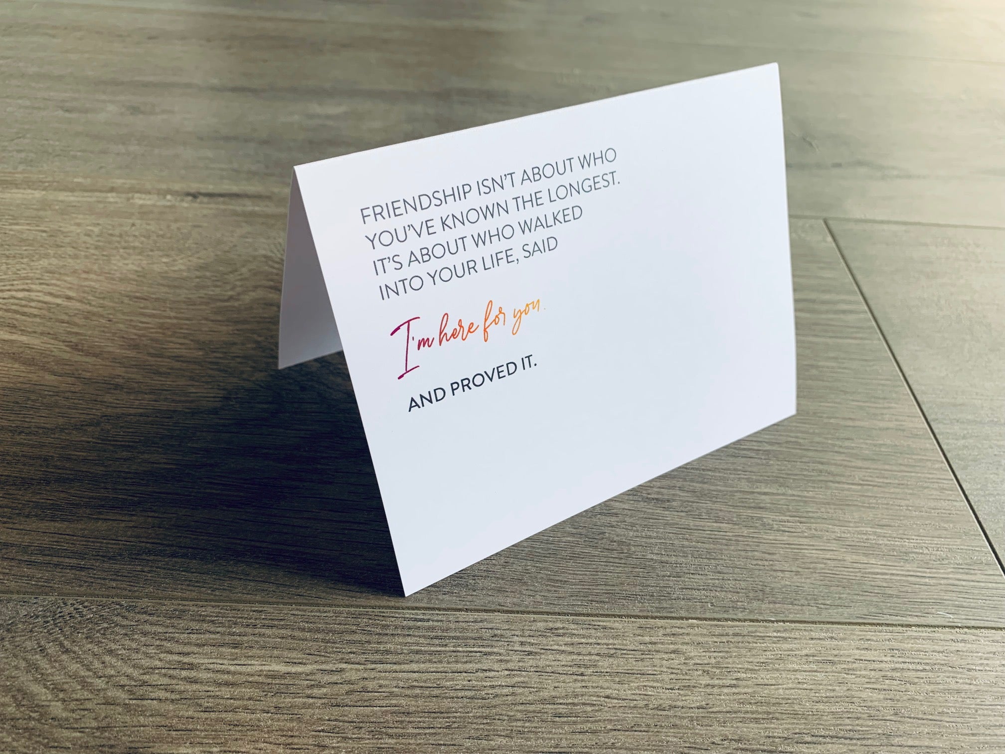 A white, folded notecard sits on a wooden floor. The card says, "Friendship isn't about who you've known the longest. It's about who walked into your life, said I'm here for you and proved it." The Friendship collection by Stationare.