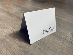 A white notecard is folded and propped up on a wooden floor. On the bottom right-hand corner, it reads, "hitched" in black ink with a heart icon dotting the I. The card is from Stationare's Bridal and Wedding collection.