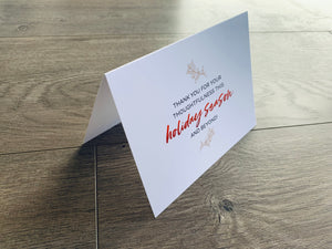 A white folded notecard sits on a wooden floor. On the front of the card, it reads, "Thank you for your thoughtfulness this holiday season and beyond!" Merry Thanks Collection by Stationare.
