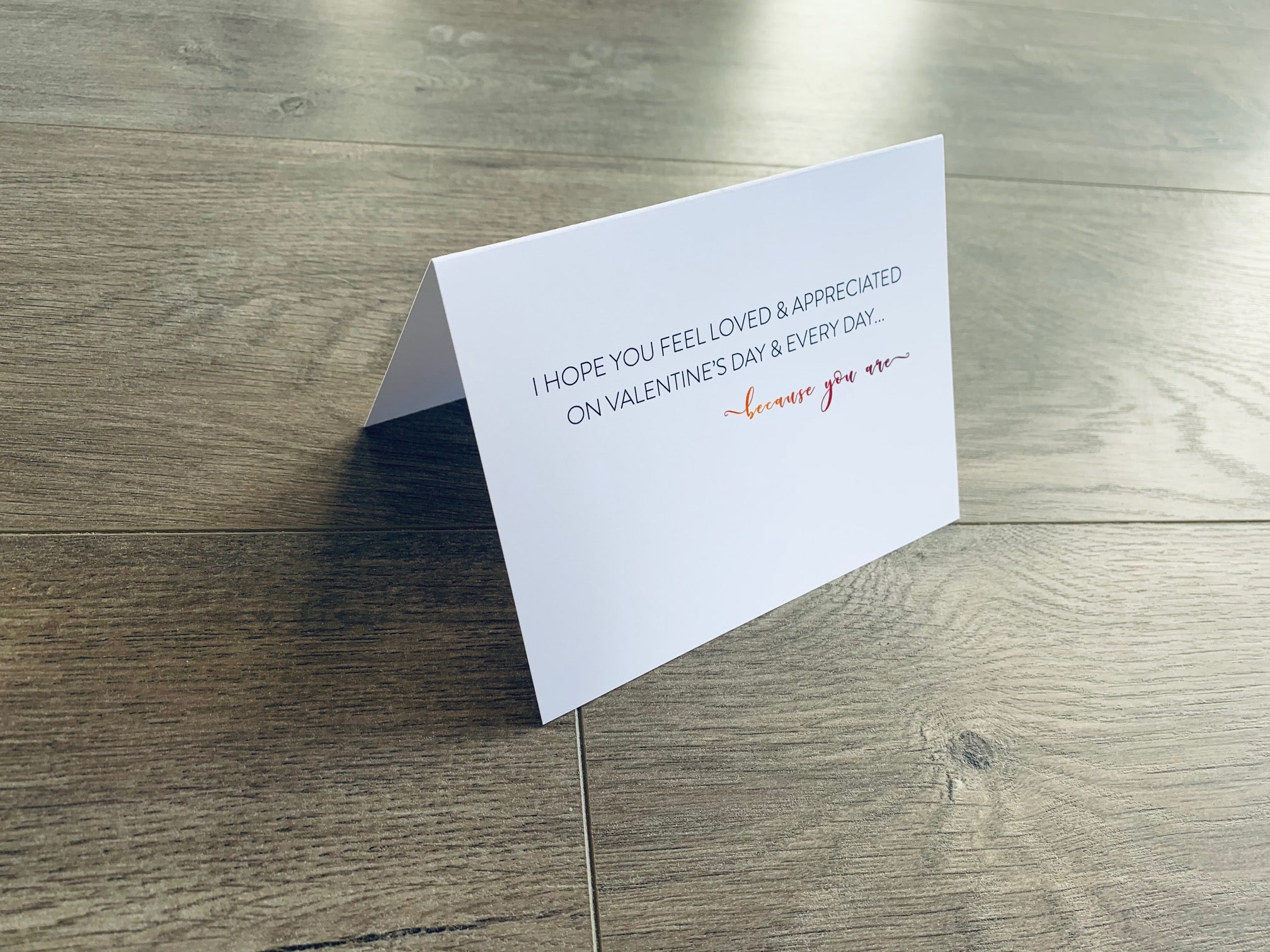 A folded white notecard is folded and propped up on a gray wooden background. The card says, "I hope you feel loved and appreciated on Valentine's Day and every day... because you are." VDay BFFs collection by Stationare.