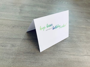 A white card is propped on a gray wood floor. The card reads, "hugs, kisses and tons of birthday wishes" birthday card by Stationare
