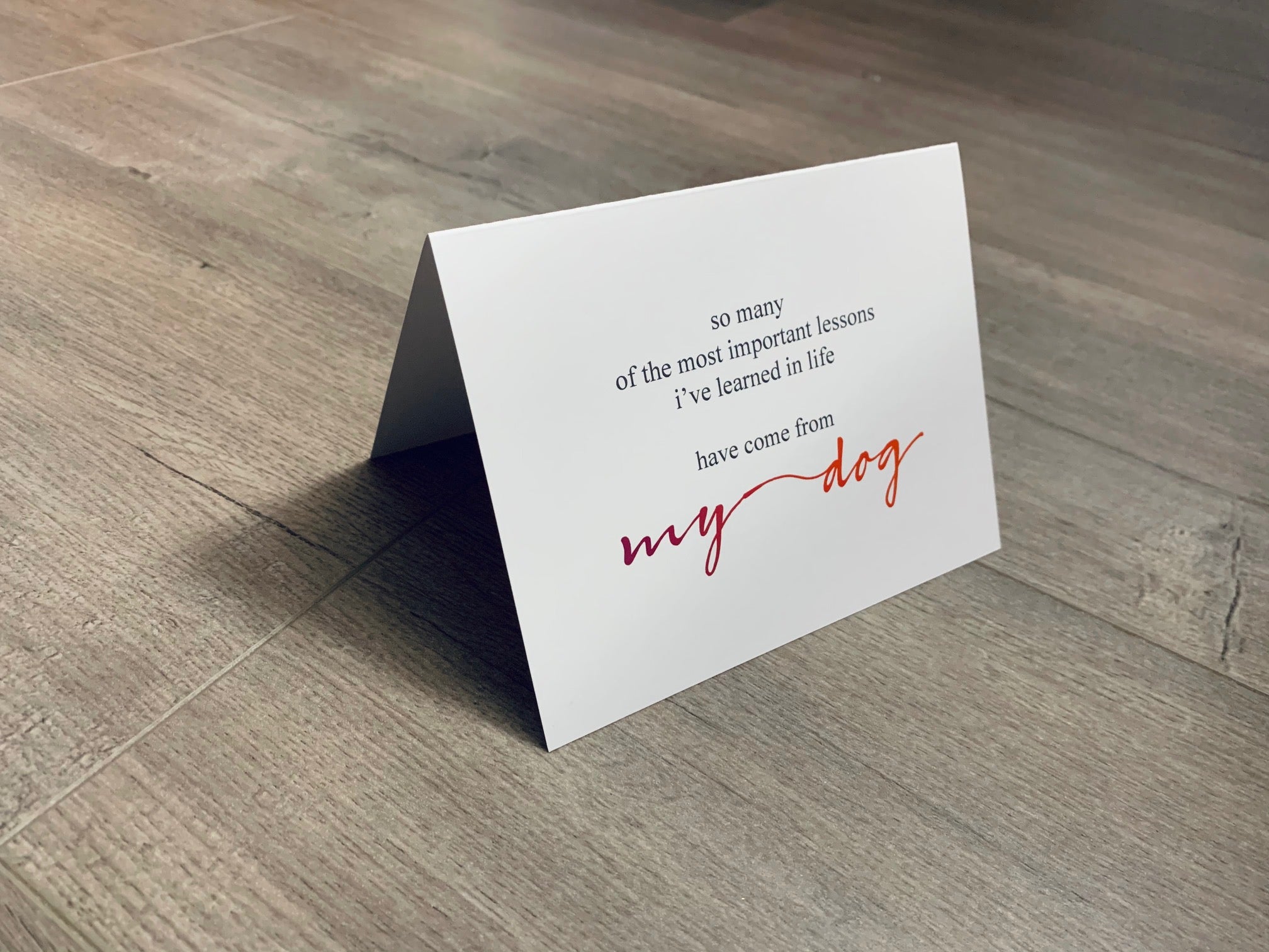 A white, folded notecard sits on a wooden floor. The card says, "so many of the most important lessons I've learned in life have come from my dog." Dog Lovers collection by Stationare.