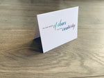 A white, folded notecard sits on a wooden floor. The card says, "In the midst of chaos, we find creativity." Creativity collection by Stationare.
