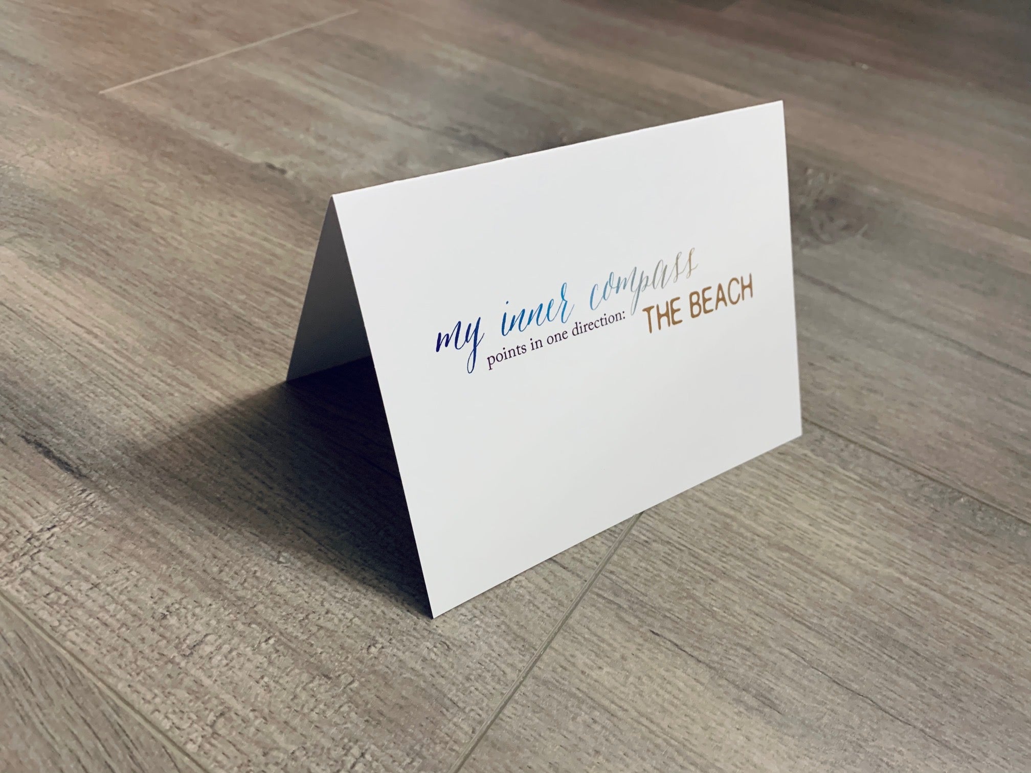 A white card is propped on a gray wood floor. The card reads, "my inner compass points in one direction - the beach." Beach Smiles Collection by Stationare.