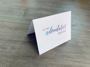 A white, folded notecard sits on a wooden floor. The card says, "Let the adventures begin!" Congrats collection by Stationare.