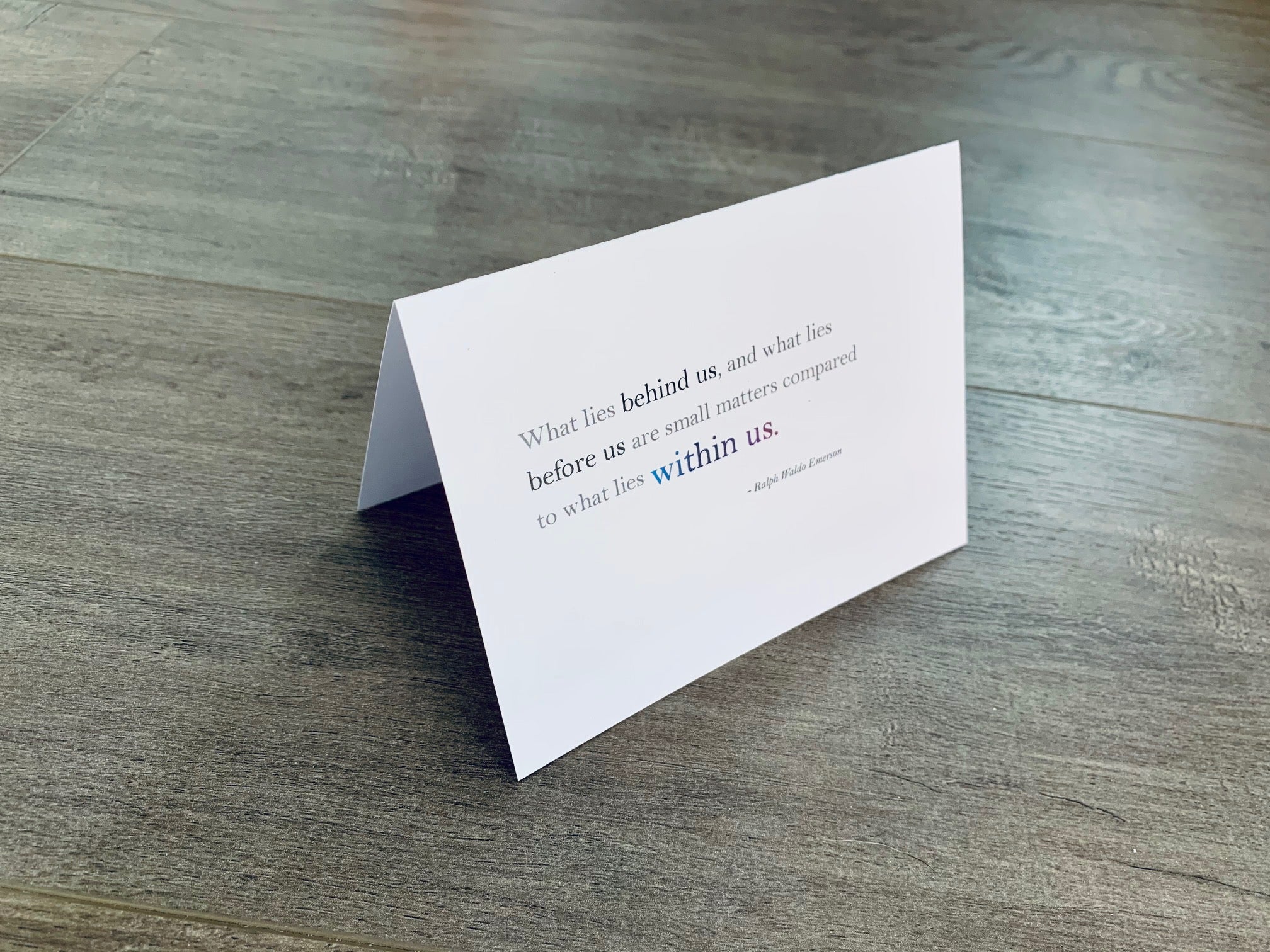 A white, folded notecard sits on a wooden floor. The card has a Ralph Waldo Emerson quote on it. It says, "What lies behind us, and what lies before us are small matters compared to what lies within us." Congrats collection by Stationare.