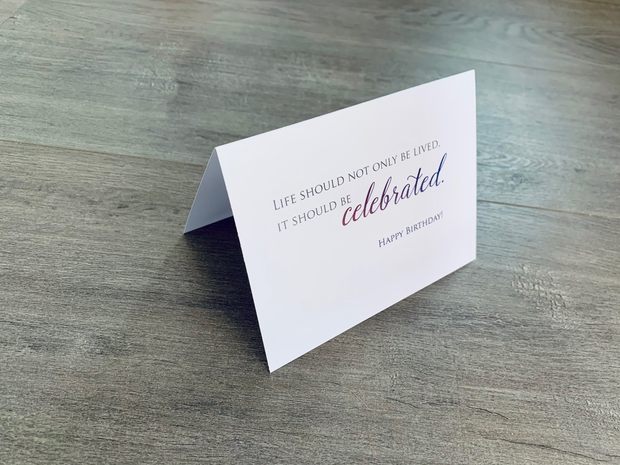 A white card is propped on a gray wood floor. The card reads, "Life should not only be lived, it should be celebrated." Birthday card by Stationare.