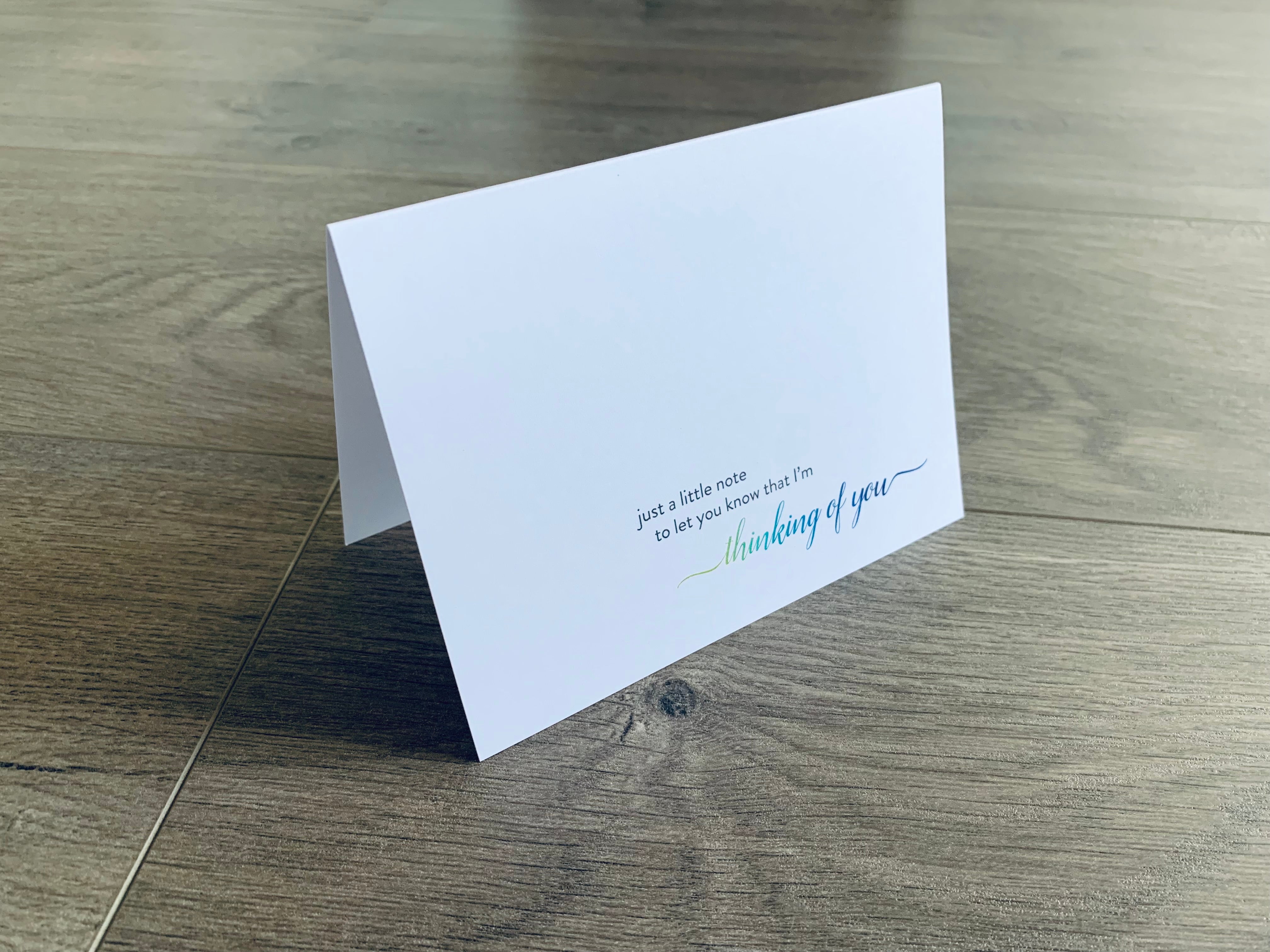 A white notecard is propped up on a gray wooden floor. The card reads, "Just a little note to let you know that I'm thinking of you." From Stationare's Thinking of You collection.
