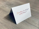A folded white notecard is propped up on a wooden floor. The card reads, "Do you believe in love at first sight or should I walk by again?" Stationare's Pick Up Lines collection.