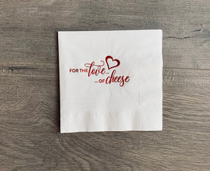 white cocktail napkin with shiny red foil. Text says - for the love of cheese - with a heart. The napkin sits on a gray wood background.