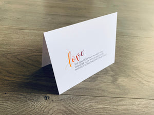 A folded white notecard is propped up on a wooden floor. The card reads, "love - the emotion that makes you wonder how you ever lived without someone or something" in a mix of block and script fonts. Love Collection by Stationare.