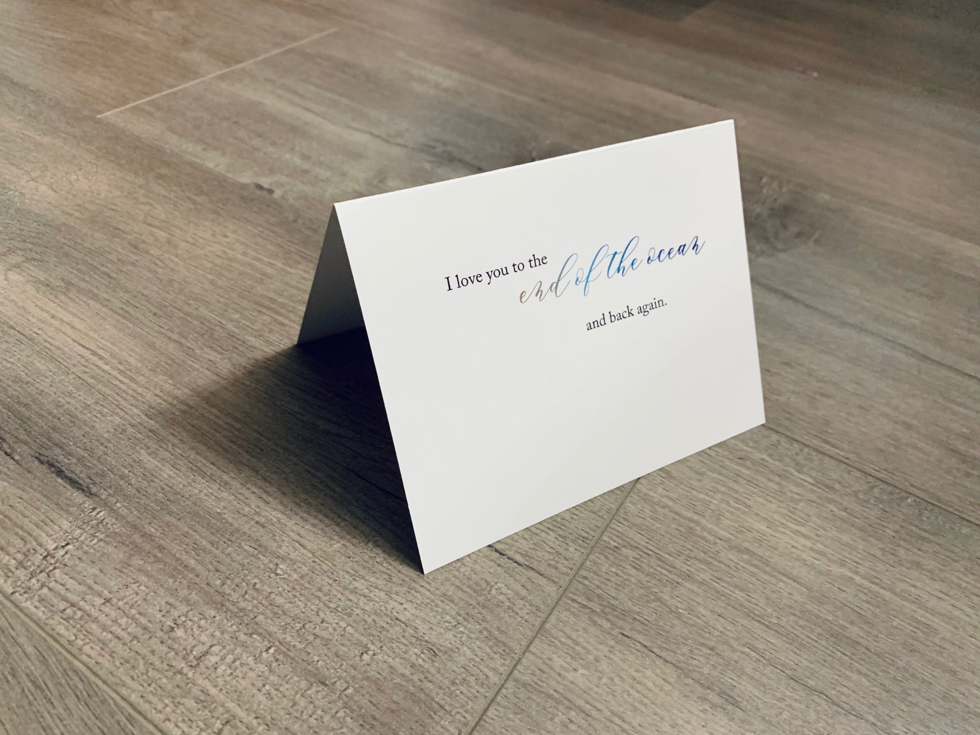 A white card is propped on a gray wood floor. The card reads, "I love you to the end of the ocean and back again." Beach Love Collection by Stationare.