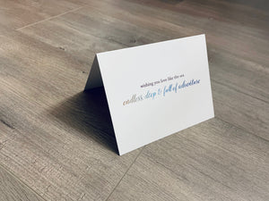 A white card is propped on a gray wood floor. The card reads, "Wishing you love like the sea... endless, deep and full of adventure." Beach Love collection by Stationare.