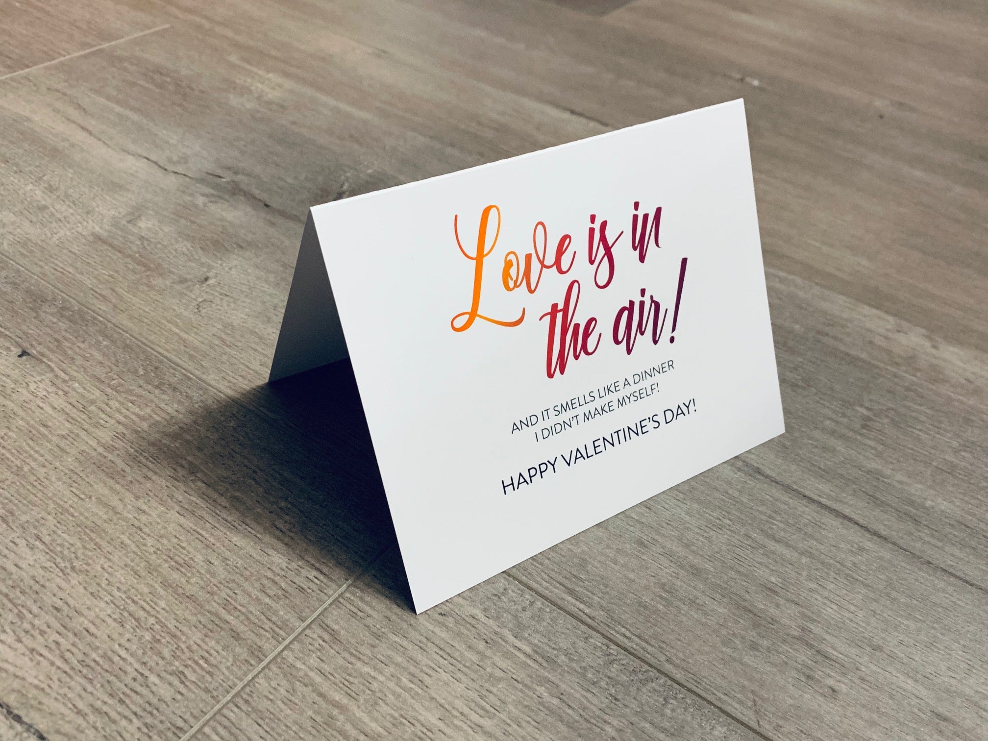 A folded white notecard is folded and propped up on a gray wooden background. The card says, "Love is in the air! And it smells like a dinner I didn't make myself! Happy Valentine's Day!" Valentine Smiles collection by Stationare.