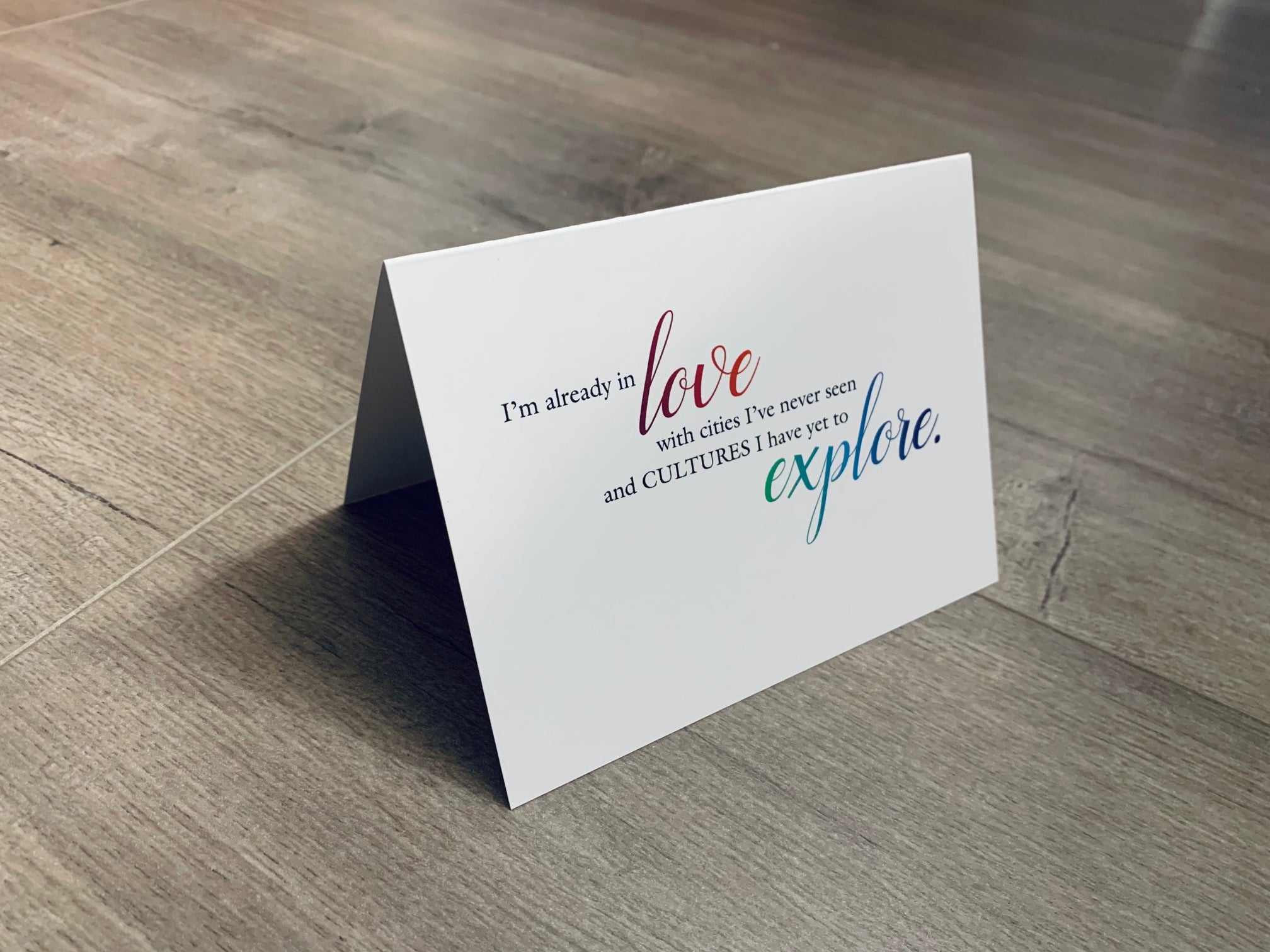 A folded white notecard is propped up on a wooden floor. The card reads, "I'm already in love with cities I've never seen and cultures I have yet to explore." Stationare's Travel Lovers collection.