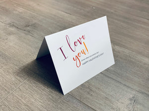 A folded white notecard is folded and propped up on a gray wooden background. The card says, "I love you! even when you annoy me. Happy Valentine's Day!" Valentine Smiles collection by Stationare.