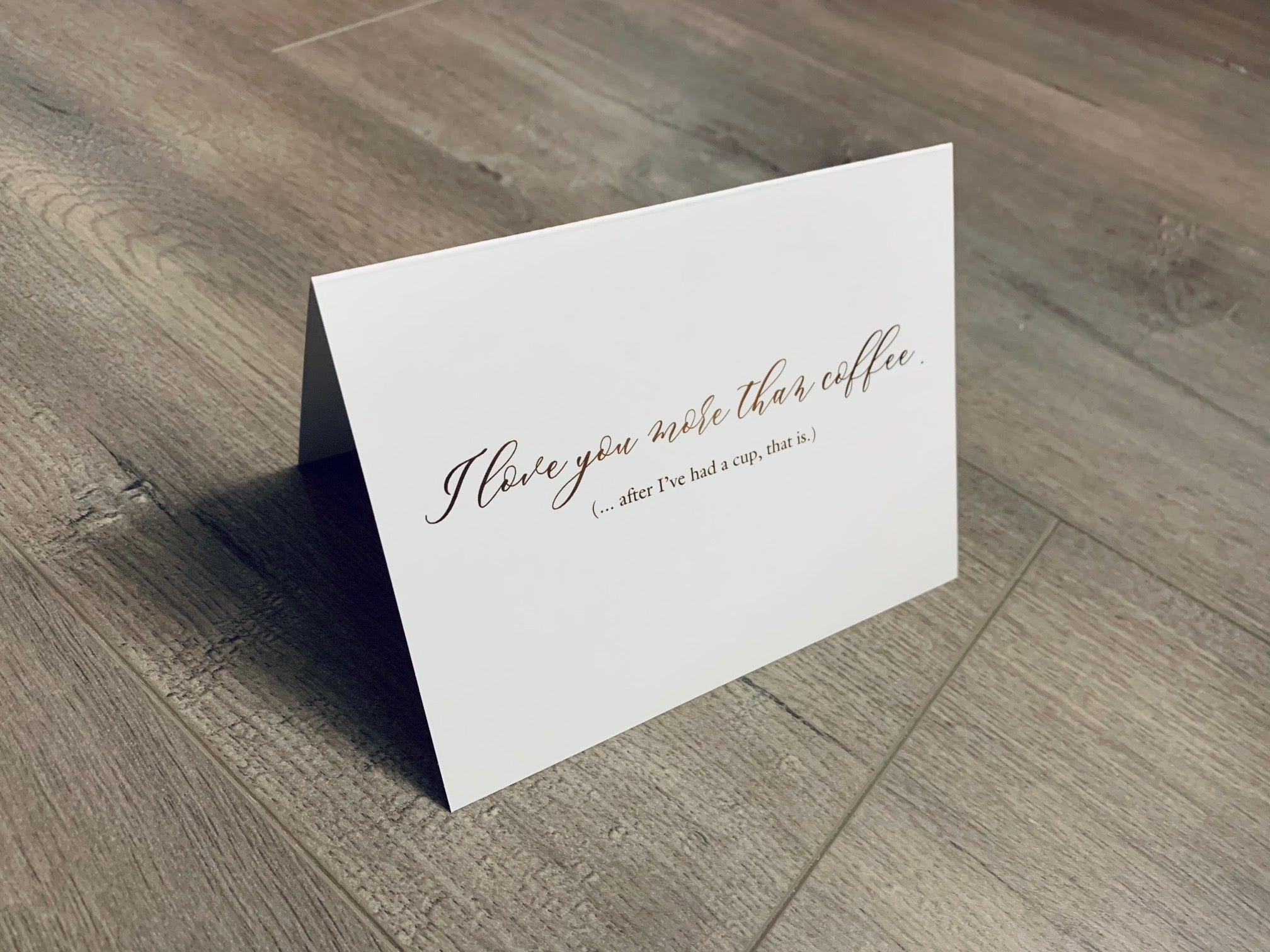 A white, folded notecard sits on a wooden floor. The card says, "I love you more than coffee. After I had a cup, that is." Coffee Lovers collection by Stationare.