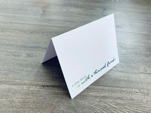 A folded white notecard is propped up on a wooden floor. The card reads, "A loyal sister is worth a thousand friends." Stationare's Sisters collection.