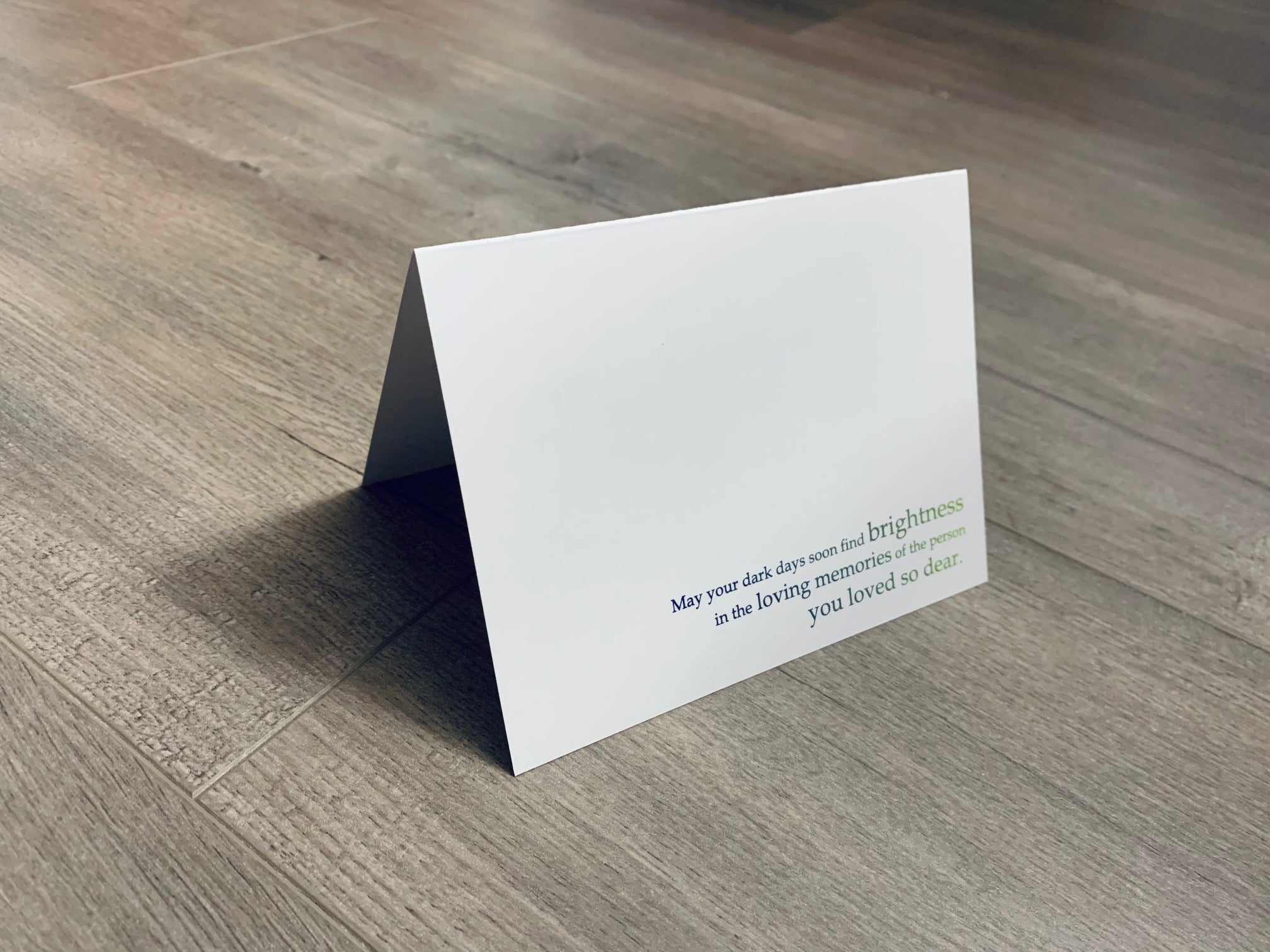 A white, folded sympathy card is folded and propped up on a gray wood floor. The card says, "May your dark days soon find brightness in the loving memories of the person you loved so dear." The printing color fades from navy to green. From the In Sympathy collection by Stationare.