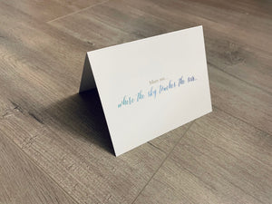 A white card is propped on a gray wood floor. The card reads, "Meet me... where the sky touches the sea..." Beach Life Collection by Stationare.