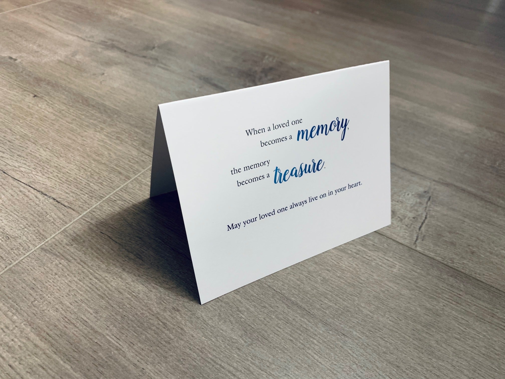 A white sympathy card is folded and propped up on a gray wooden floor. The card says, "When a loved one becomes a memory, the memory becomes a treasure. May your loved one always live on in your heart." In Sympathy Collection by Stationare.