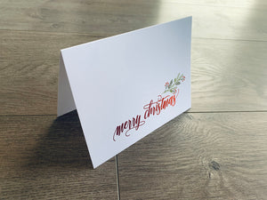 A white folded notecard is propped up on a gray wooden floor. The card reads, "Merry Christmas," in a red script font and is accented with a branch of holly.