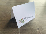 A white folded notecard is propped up on a gray wooden floor. The card reads, "Merry Christmas," in a black script font and is accented with a branch of holly.