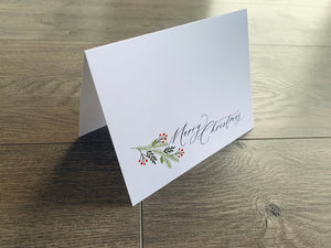 A white folded notecard is propped up on a gray wooden floor. The card reads, "Merry Christmas," in a black script font and is accented with a branch of holly.
