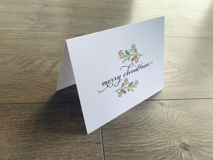 A white folded notecard is propped up on a gray wooden floor. The card reads, "Merry Christmas," in a black script font and is accented with a branch of holly.