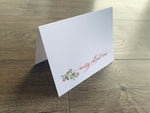 A white folded notecard is propped up on a gray wooden floor. The card reads, "Merry Christmas," in a red script font and is accented with a branch of holly.