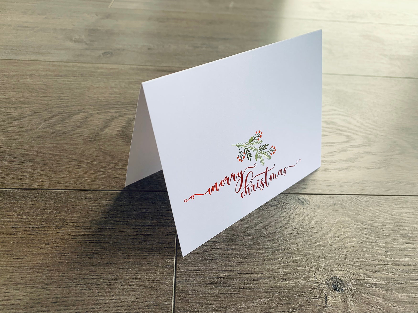 A white folded notecard is propped up on a gray wooden floor. The card reads, "Merry Christmas," in a red script font and is accented with a branch of holly.