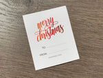 A rectangular white sticker gift tag lies on a wooden background. The words "merry christmas" are written in a bold script font that ranges from orange to dark red. Below the sentiment are the words "to" and "from" for the gifter hand-write. Merry Christmas Sticker gift tag by Stationare.