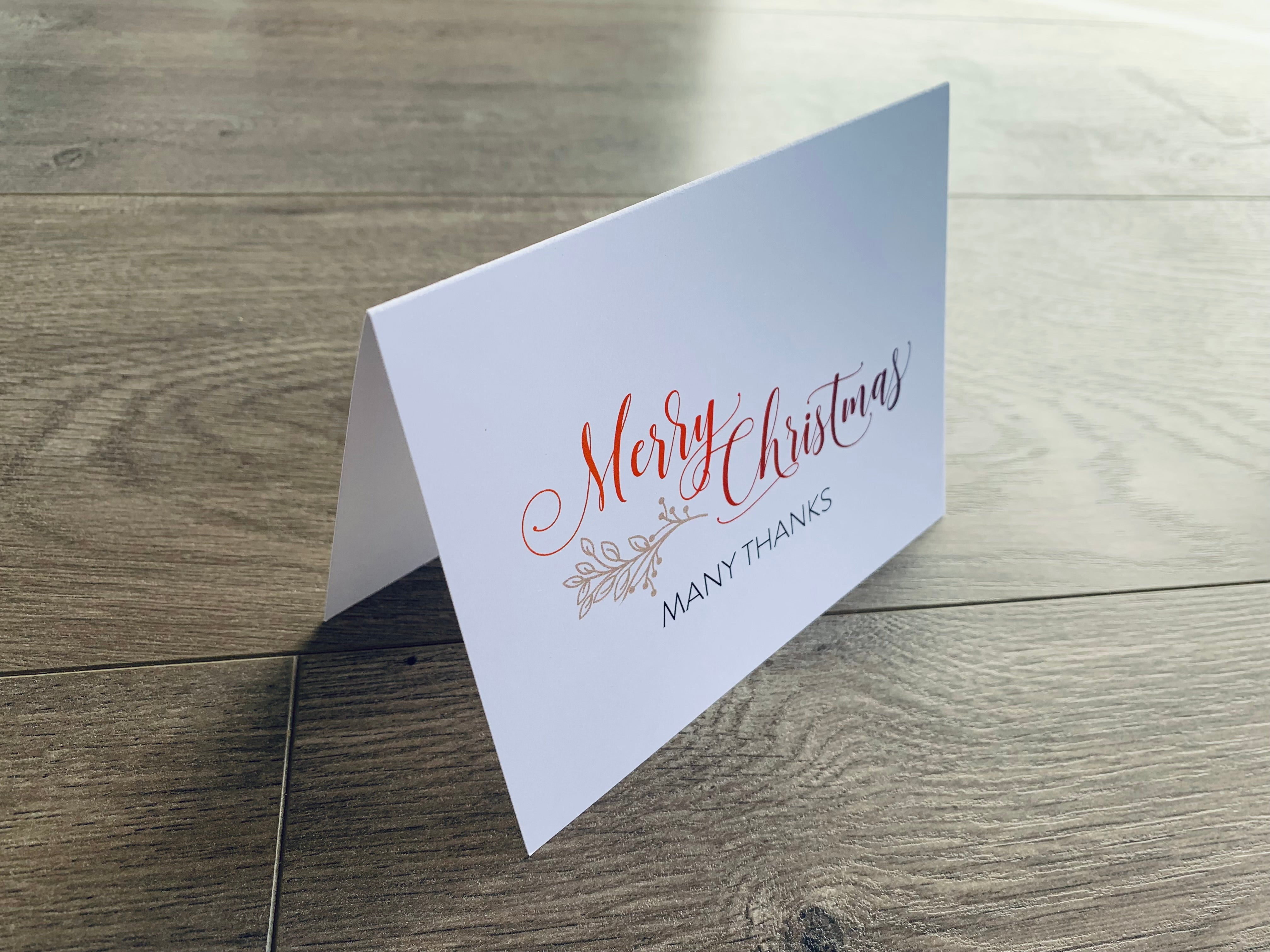 A white folded notecard sits on a wooden floor. On the front of the card, it reads, "Merry Christmas. Many Thanks." Merry Thanks Collection by Stationare.
